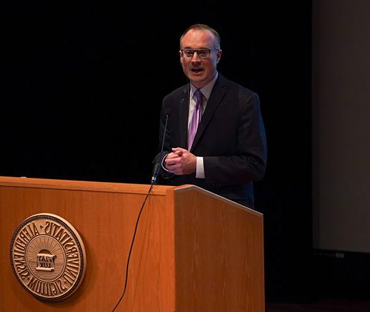 man with coat and tie, glasses, speaking at podium